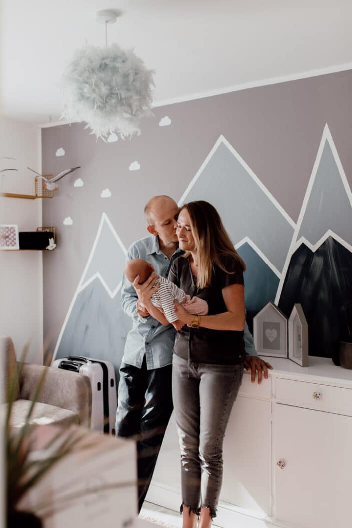 Mum and dad are standing in newborn baby room and dad is kissing mum. Mum is holding newborn baby girl. They are standing in a lovely newborn baby room with mountain painted on the wall. Newborn photographer in Hampshire. Ewa Jones Photography