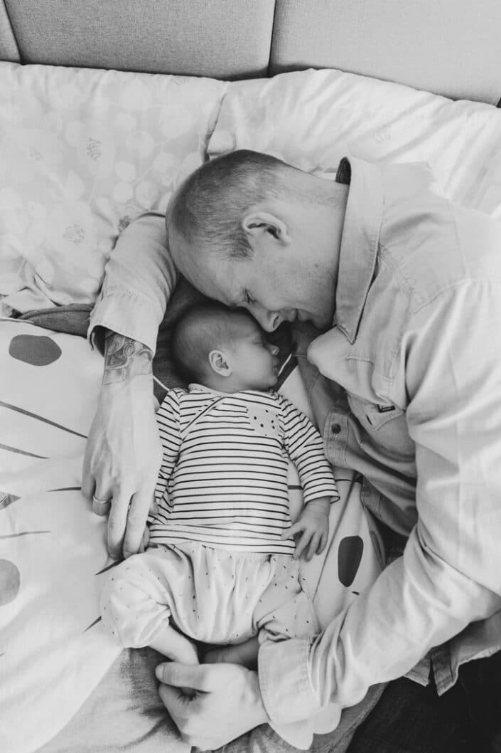 Dad is laying on bed and has his arm wrapped around his baby girl. Baby girl is sleeping. Newborn photography in Hampshire. Newborn baby feet detail shoot. Newborn baby is holding his feet together whilst laying on the bed. Newborn baby photography in Hampshire. Ewa Jones Photography