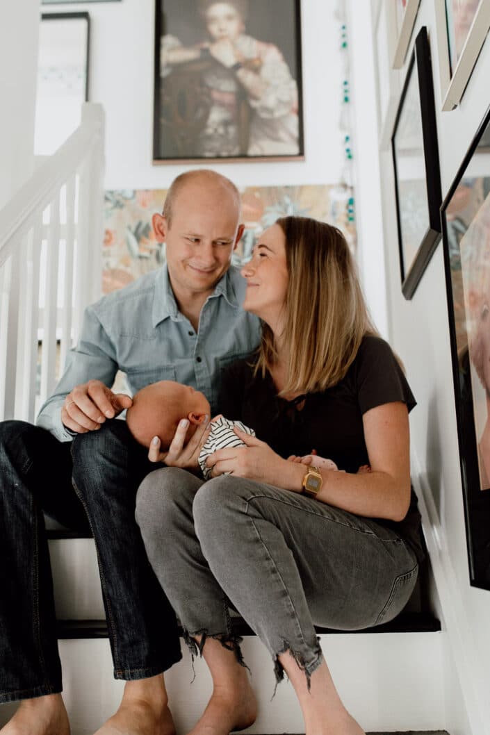 Mum and dad are sitting on the stairs and lovingly looking at each other. Mum is holding newborn baby. Newborn photographer in Hampshire. Ewa Jones Photography