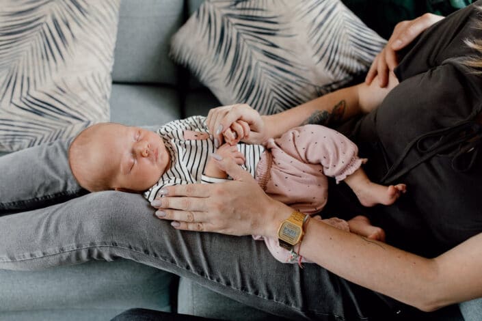 Mum is sitting on the sofa and holding her newborn baby girl on her leg. Baby girl is sleeping and she is wearing a stripy top and pink trousers. In-home newborn baby photo session in Basingstoke, Hampshire. Newborn baby feet detail shoot. Newborn baby is holding his feet together whilst laying on the bed. Newborn baby photography in Hampshire. Ewa Jones Photography