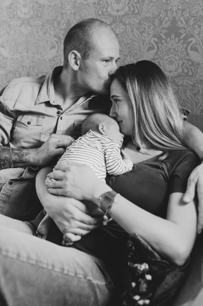 Mum and dad are sitting on the sofa and dad has his hand wrapped around mum. Mum is holding her newborn baby girl and baby girl is sleeping. Newborn photography in Hampshire. Ewa Jones Photography