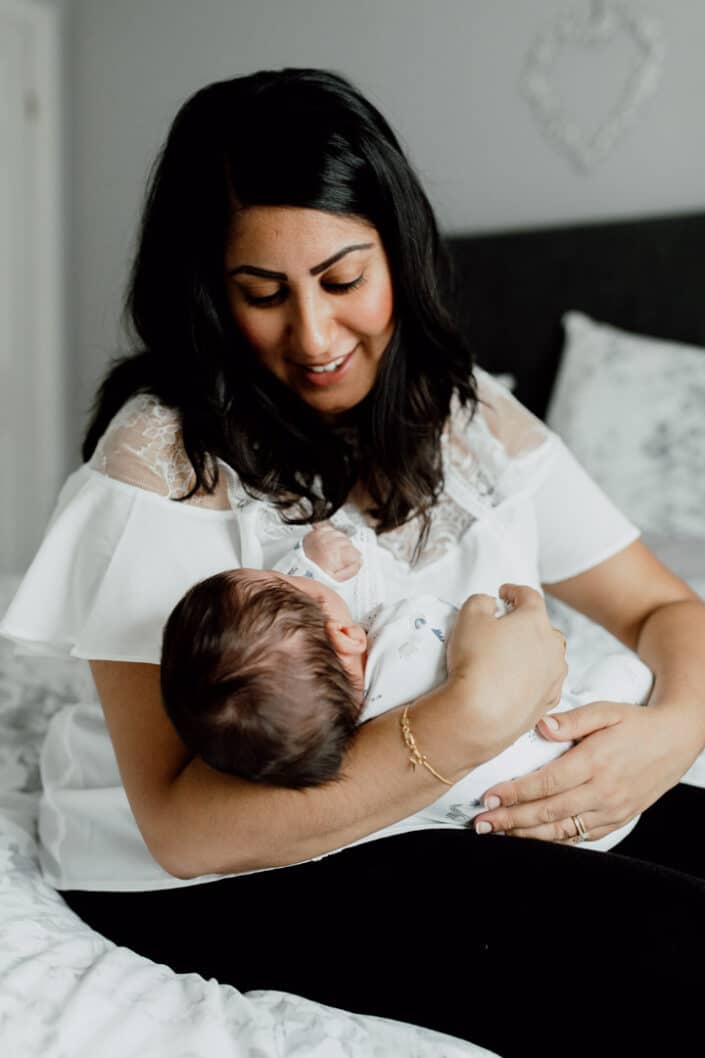 Mum is sitting at the edge of the bed and looking down at her newborn baby boy. Mum is wearing lovely white top and baby boy is wearing white baby grow. Newborn photographer in Fleet, Hampshire. Ewa Jones Photography