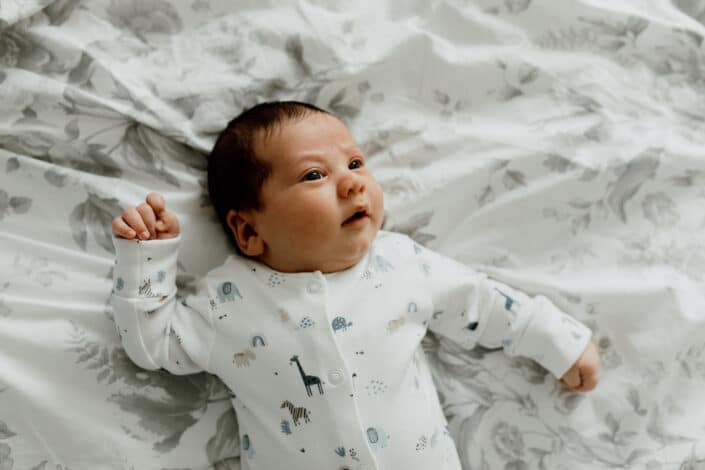 Newborn baby is laying on bed and looking up. he is wearing lovely white baby grow with animal prints. in-home lifestyle newborn baby photography in Hampshire. Ewa Jones Photography