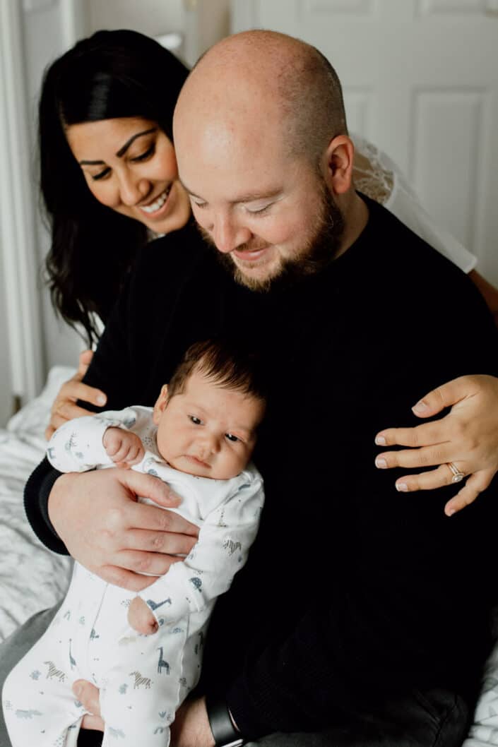 Mum and dad are on the bed and mum is holding dad lovingly. Dad is holding his newborn baby boy. Newborn baby photography in Hampshire. Ewa Jones Photography