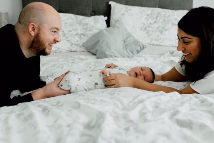 Newborn baby boy is laying on bed and mum and dad are kneeling on the side of the bed. Both are holding baby's feet and hands. Newborn photography in Hampshire. Newborn baby feet detail shoot. Newborn baby is holding his feet together whilst laying on the bed. Newborn baby photography in Hampshire. Ewa Jones Photography