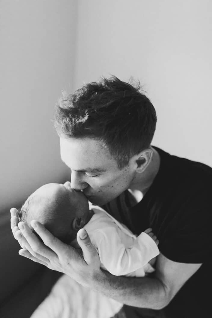 Dad is sitting on the bed and kissing his newborn baby. Black and white photography. Hampshire newborn photoshoot. Ewa Jones Photography