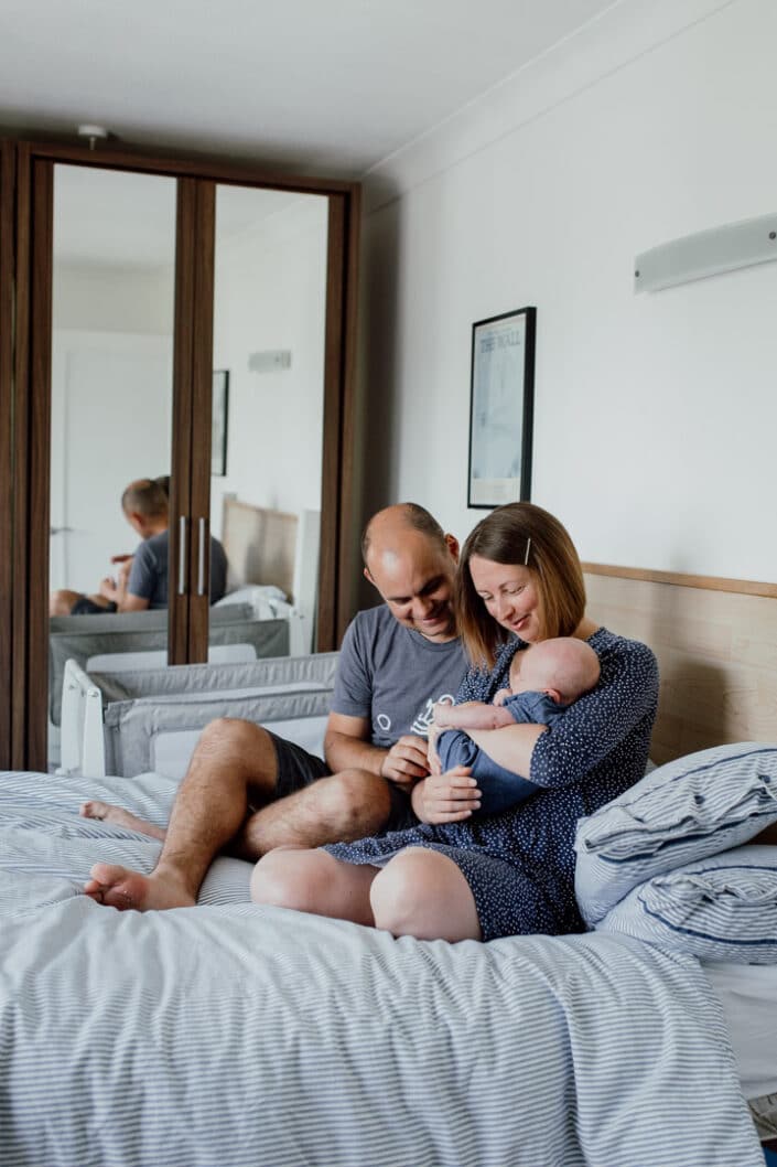 Mum and dad are sitting on the bed and looking at their baby. Mum is holding baby and both parents are smiling. Newborn baby photography in Basingstoke. Hampshire. Ewa Jones Photography