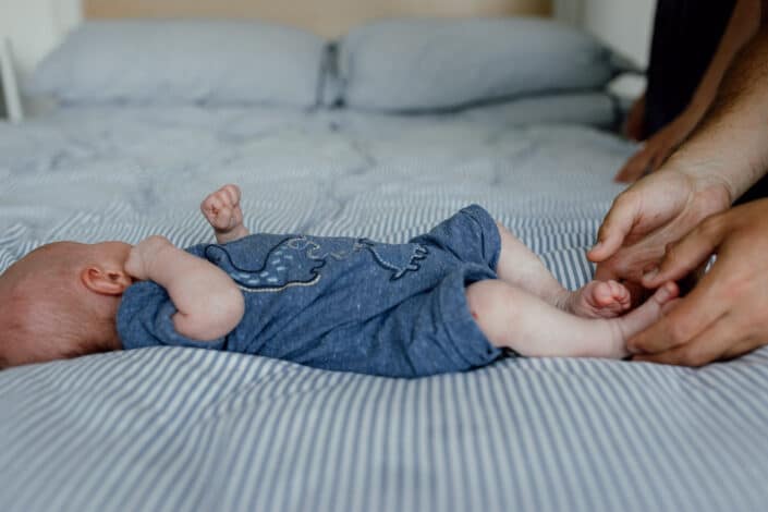 Baby is laying on bed and dad is holding newborn baby feet. In-home lifestyle newborn baby photography in Hampshire. Close up detail shoot of newborn baby feet. and his mum is holding his feet. Newborn baby photographer in Fleet, Hampshire. Ewa Jones Photography