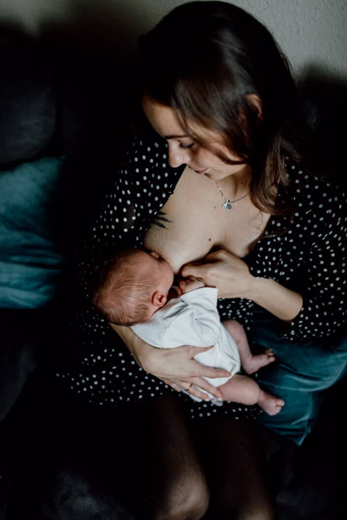 Mum is sitting on the sofa and looking down at her newborn baby. She is breastfeeding her baby. Mum is wearing black dress with white dots. Newborn photographer in Hampshire. Ewa Jones Photography