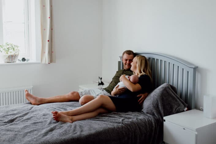 Mum and dad sitting on the sofa with a newborn baby looking at each other and smiling. Natural newborn baby photoshoot in Basingstoke, Hampshire. Ewa Jones Photography