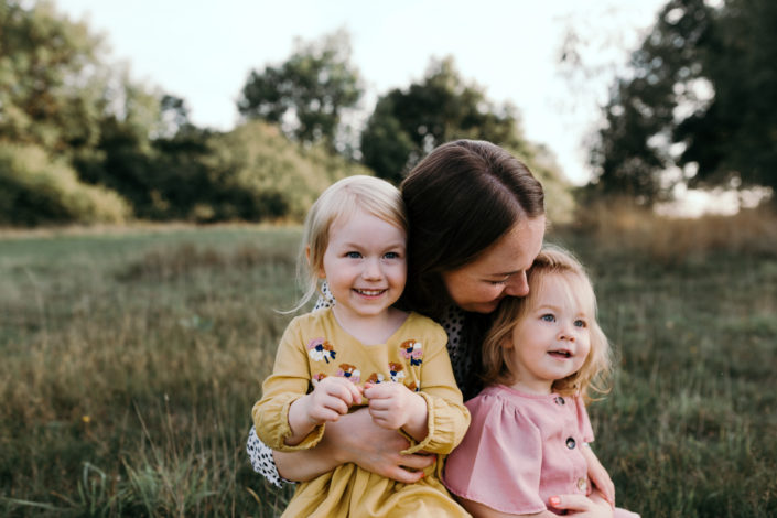Mum and two daughters are sitting on the grass cuddling each other. Happy family moments. Family photoshoot in Basingstoke. Ewa Jones Photograpy