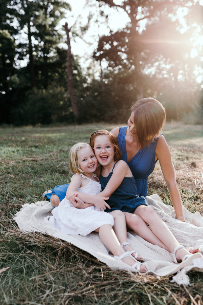 Mum sitting on the blanket with two girls. One girl is wearing blue dress and one is wearing a white dress. Mum is looking at the girls and they are laughing. Hampshire family photography. Ewa Jones Photography