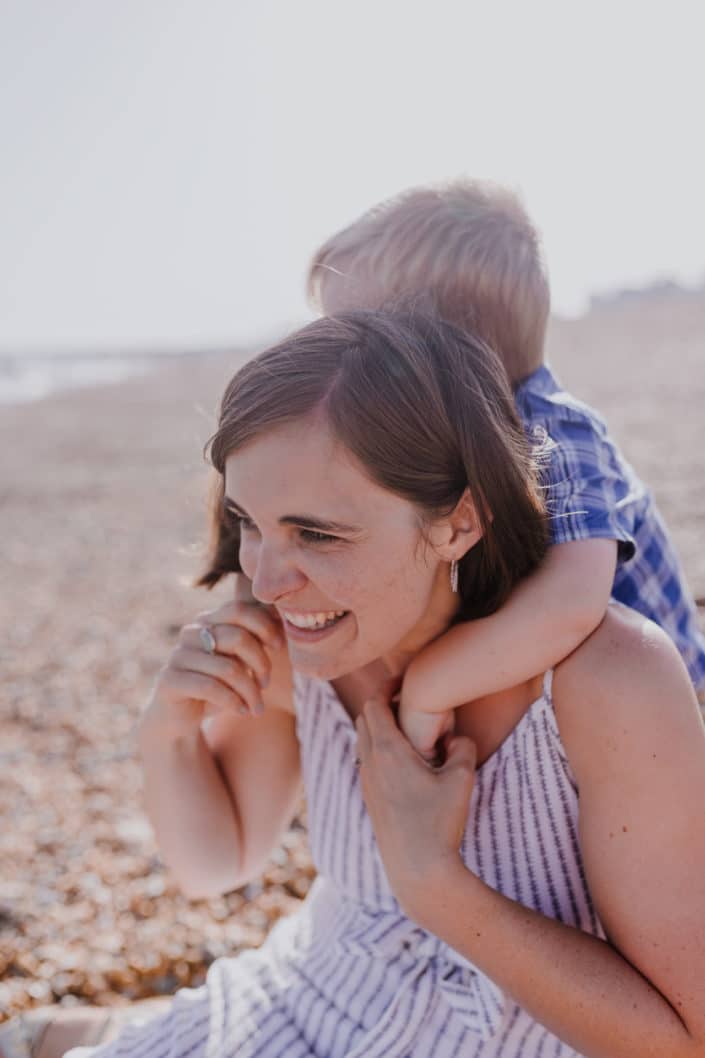 Mum is sitting on the beach and laughing. Her little boy has his arms wrapped around his mum neck. Lovely mother and son connection. Family lifestyle photography in Hampshire. Ewa Jones Photography