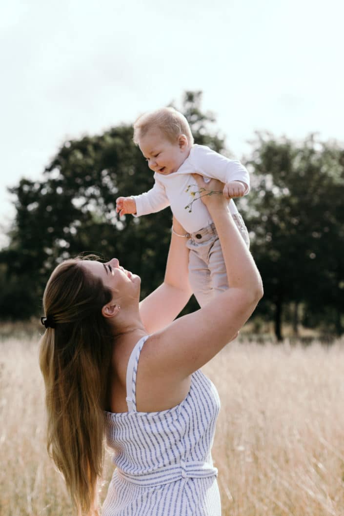 Mum is holding in the air her little boy. Boy is smiling to mum. They are in the fields and the sun is shining. Happy mother and child photosession. Family photography in Hampshire. Ewa Jones Photography