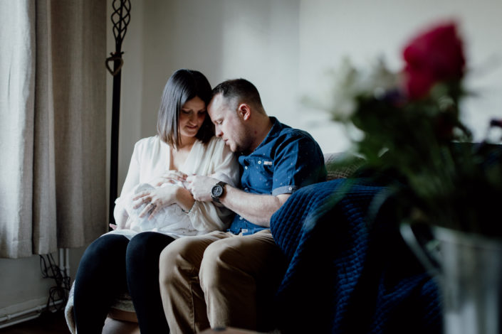 Mum and dad are sitting on the sofa and looking lovingly at their newborn baby. Lifestyle newborn photography in Basingstoke. Ewa Jones Photography