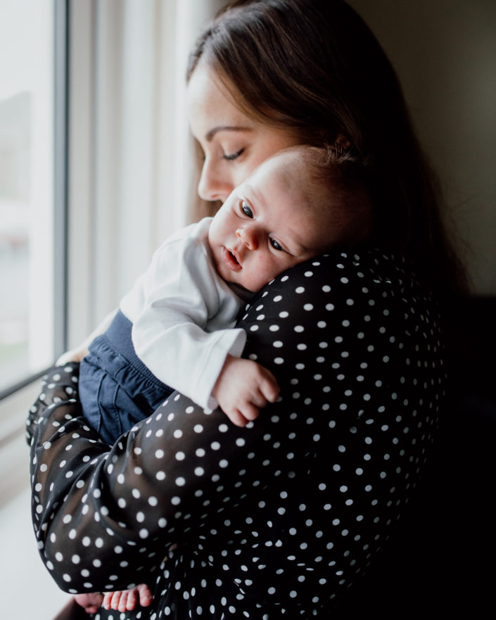Mum holding newborn baby and standing next to the window. Mum is wearing black dress with white dots. Newborn photography in Hampshire. Ewa Jones Photography