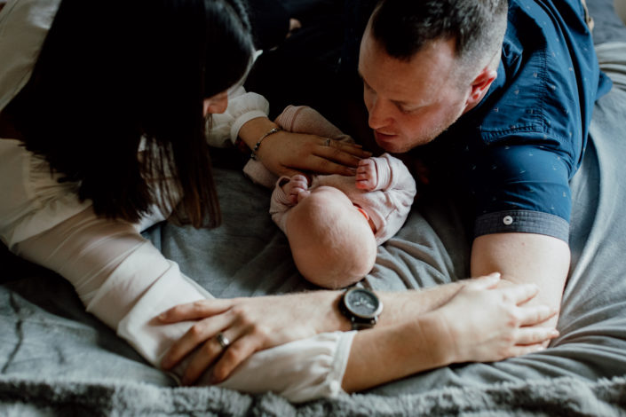 Mum and dad are laying on the bed. Newborn baby is laying in the middle. Mum and dad are holding hands. Natural lifestyle newborn photoshoot in Basingstoke. Ewa Jones Photography