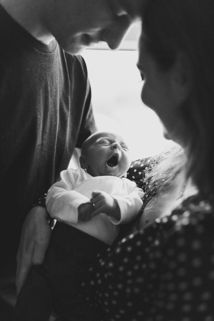 Mum and dad are holding a newborn baby and standing in front of the window. Black and white family photograph. Hampshire newborn photoshoot. Ewa Jones Photography