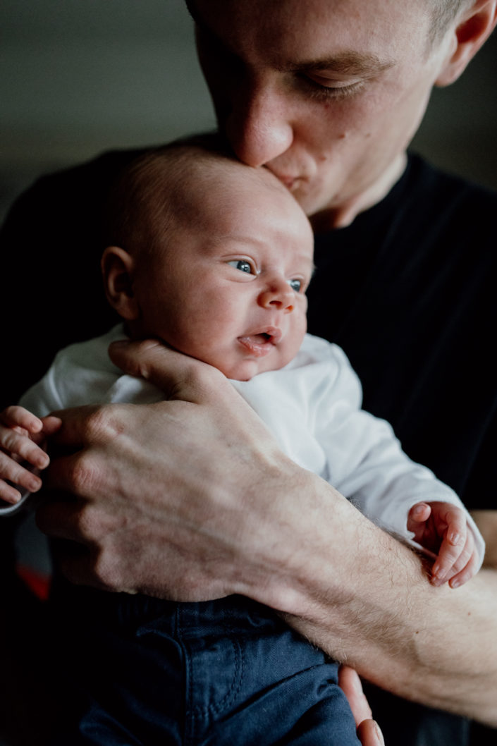Dad is holding newborn baby and standing next to the window. dad is wearing black t-shirt. Newborn photography in Hampshire. Ewa Jones Photography