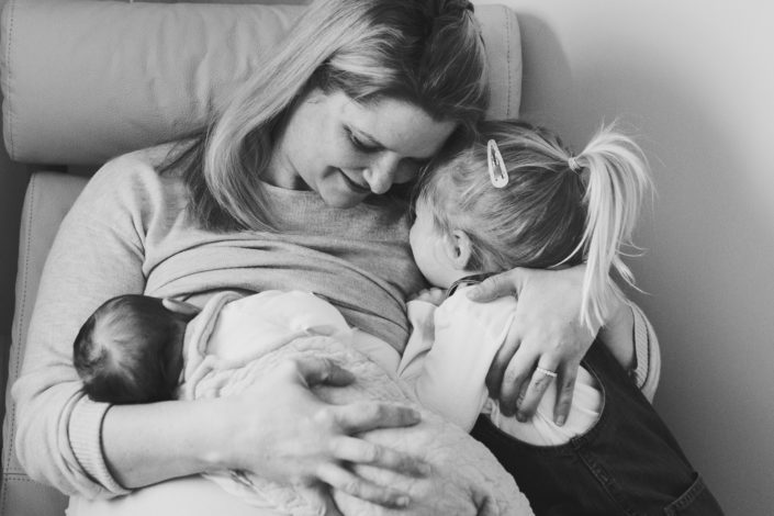 Mum is sitting on the chair and breastfeeding newborn baby. Mum is at the same time cuddling her older daughter. Newborn lifestyle family photography in Hampshire. Ewa Jones Photography