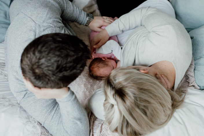 Mum and dad are laying on the bed and mum is breastfeeding her newborn baby. Both parents are looking at the baby. Natural newborn photoshoot in Hampshire. Ewa Jones Photography