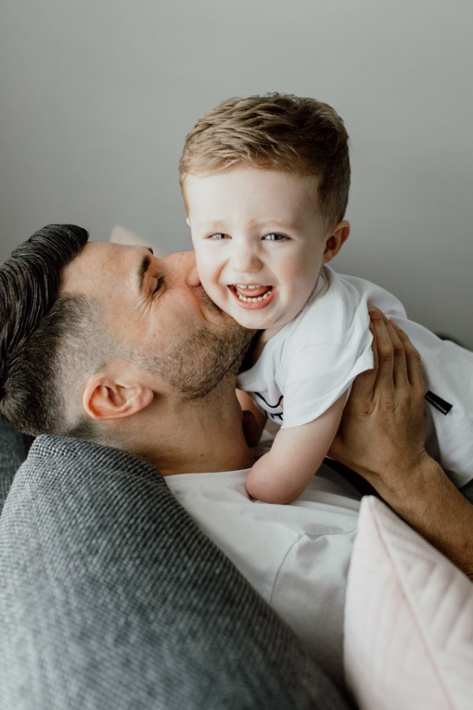 Dad is sitting on the sofa and kissing his toddler boy. Boy is looking at the camera and smiling. Favourite images from a newborn session. family photographer in Basingstoke. Ewa Jones Photography