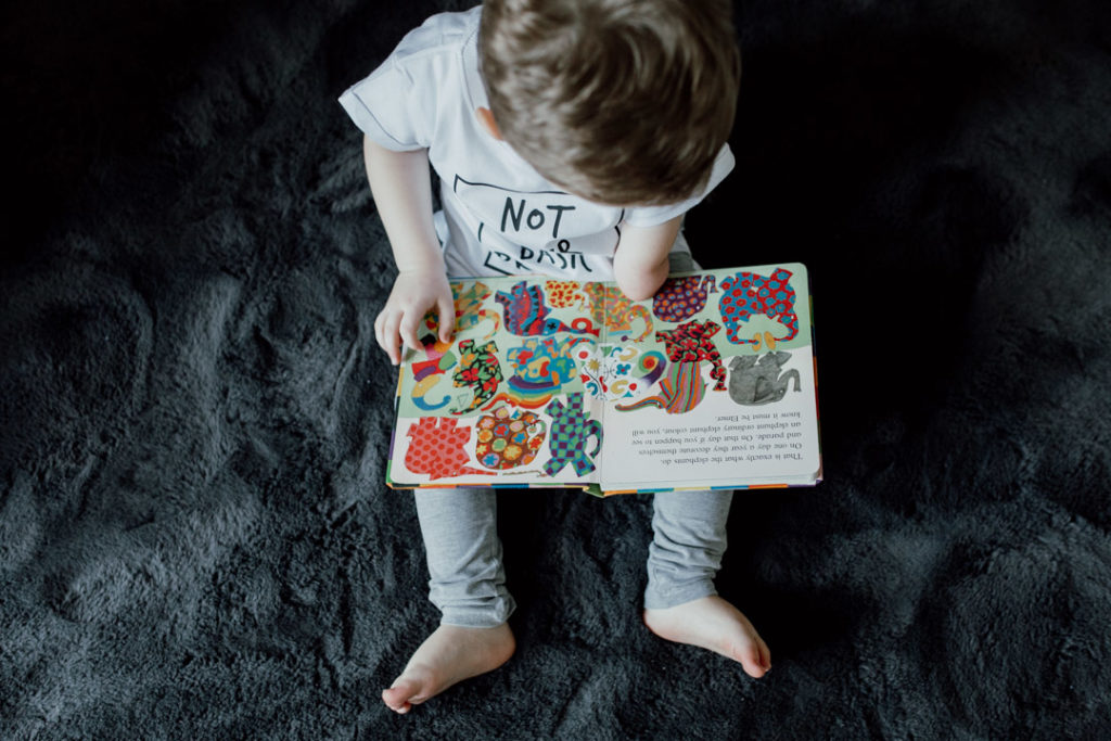 Boy is sitting on the floor and reading an Elmer book. Boy is looking down at the colorful pictures in the book. Family lifestyle photography in Basingstoke. Ewa Jones Photography