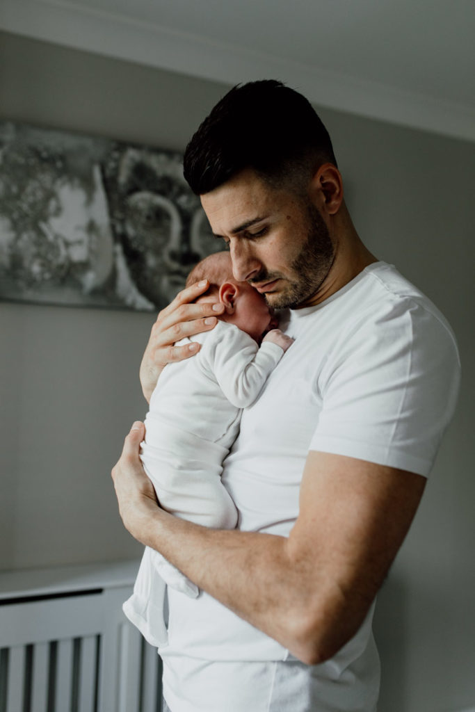 Dad is standing and cuddling his newborn baby. Dad is wearing white shirt and baby is wearing a white baby grow. family photographer in Basingstoke. Favourite images from a newborn session. Ewa Jones Photography