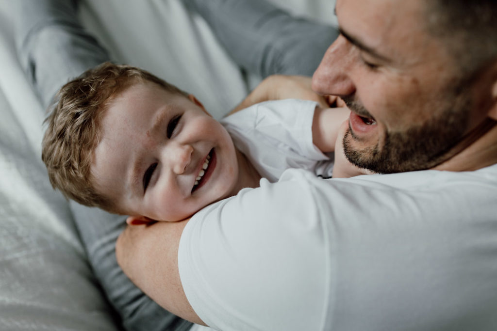 Dad and his toddler son are playing tickles on the bed, Toddler boy is looking at the camera and smiling. Family lifestyle photography in Basingstoke