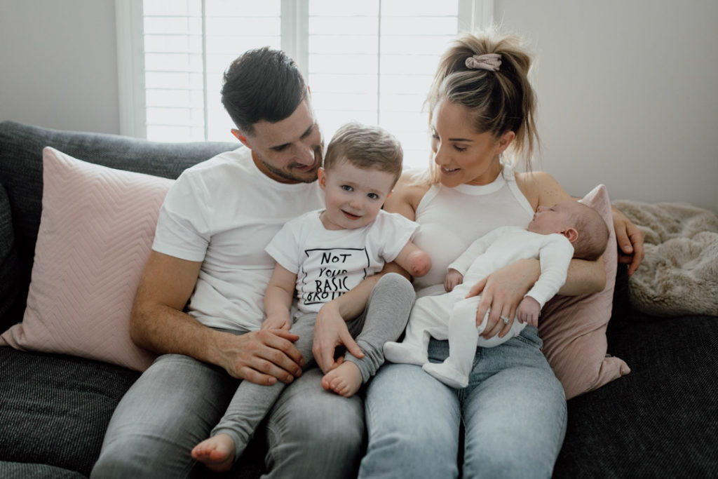 Mum and dad are sitting on the sofa. Older boy is sitting on daddy's lap and mum is holding a newborn baby. Both mum and dad are looking at the older boy. They all wear white t-shirts and light jeans. Family photographer in Basingstoke. Ewa Jones Photography