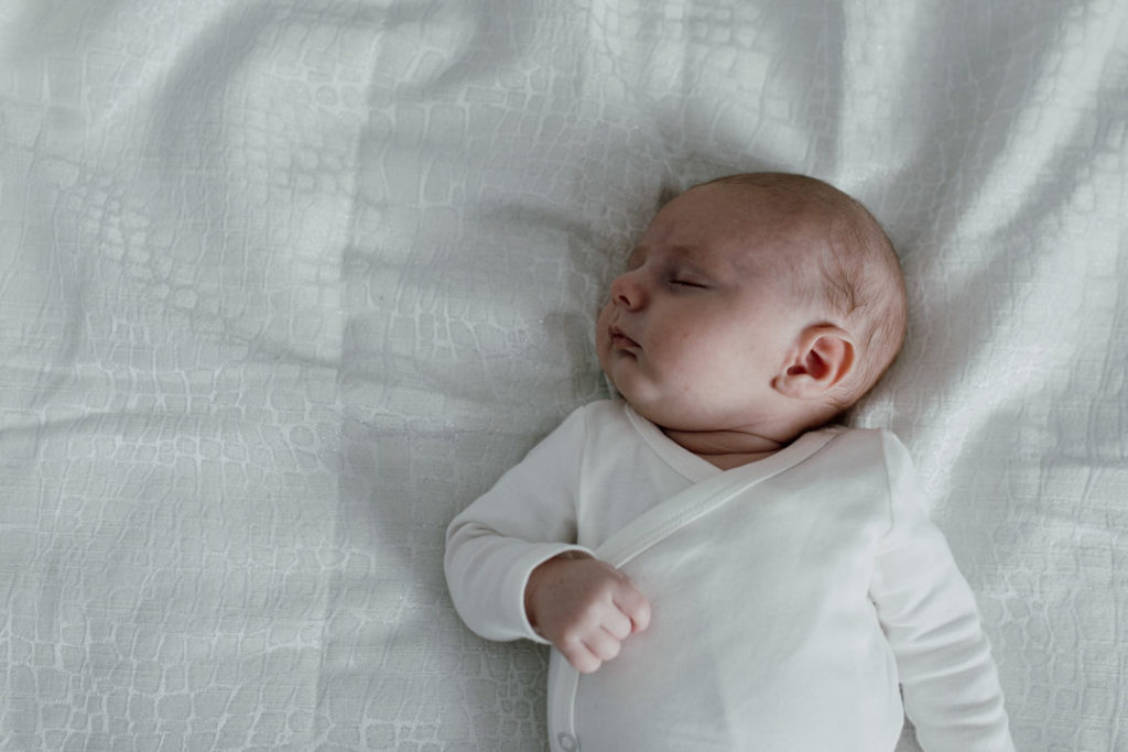 Newborn baby is laying on bed and he is sleeping. Baby is wearing white baby grow and laying on white bed sheets. Newborn photographer in Basingstoke. Favourite images from a newborn session. Ewa Jones Photography