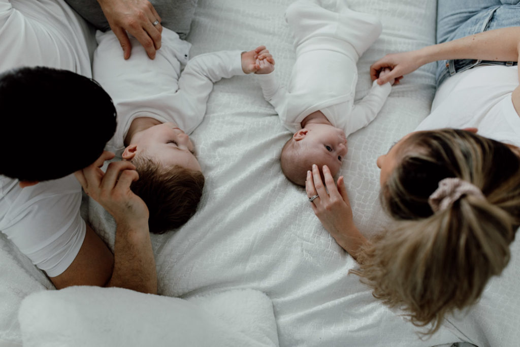 Mum and dad and two boys are laying on the bed. Shoot is taken from the top. Mum is touching newborn baby head and dad is looking at the toddler boy. Family lifestyle photography in Basingstoke. Favourite images from a newborn session. Ewa Jones Photography