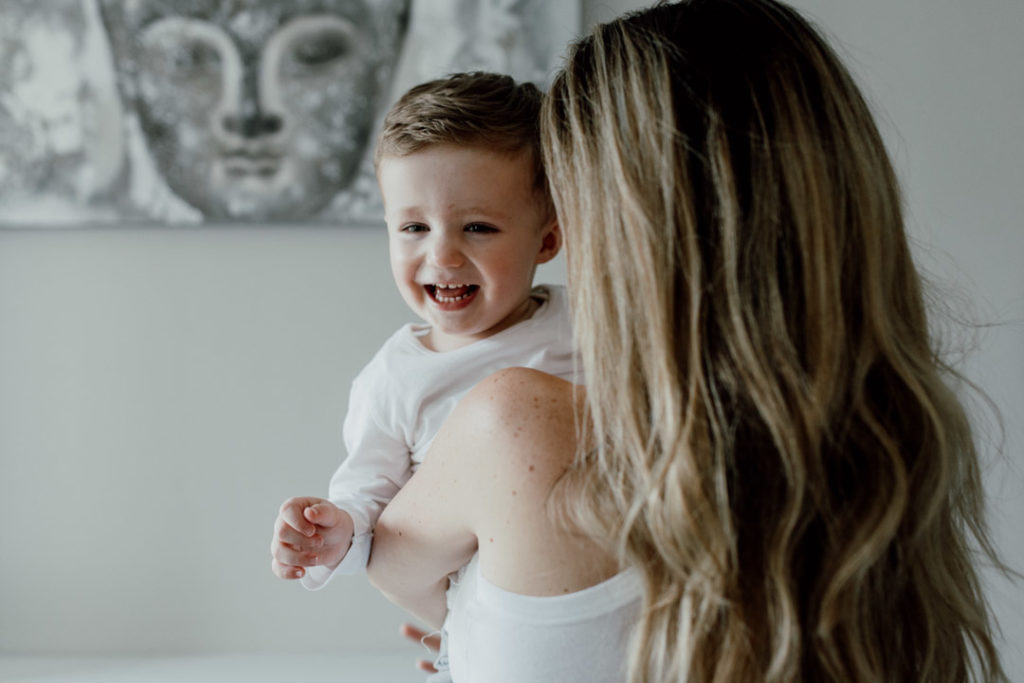 Mum is holding her toddler boy. Her head is facing a boy and boy is laughing to the camera. boy and mum are wearing white tops. Favourite images from a newborn session. Family lifestyle photography in Basingstoke. Ewa Jones Photography