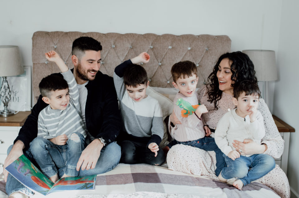 Family of six is sitting on the bed and looking at each other. Two boys are raising their hands and mum and dad are lovingly watching them. Family photography in Hampshire. Ewa Jones Photography