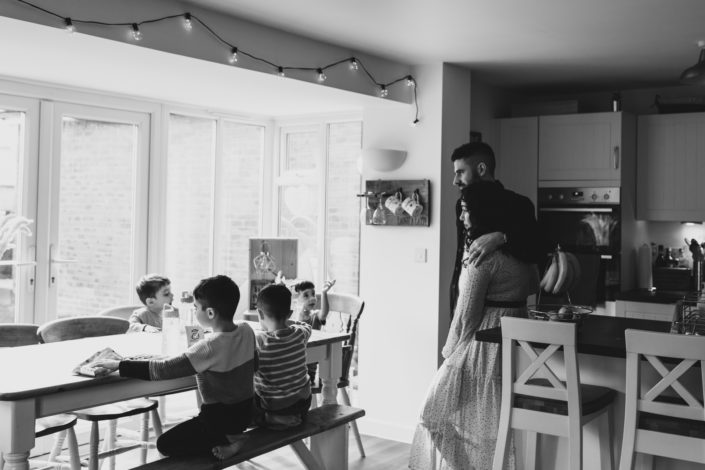 Mum and dad are standing in the kitchen looking at their boys eating lunch. Boys are sitting on the bench and eating. Candid family moment. Family photographer ins Hampshire. Ewa Jones Photography