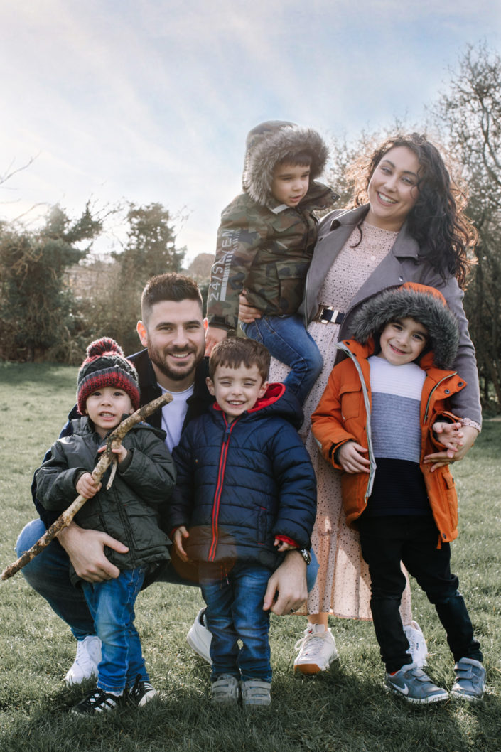 Family portrait in the park. Mum is standing and holding her son. Dad is squatting down and holding two boys. Oldest boy is on mum side. All smiling. Ewa Jones Photography