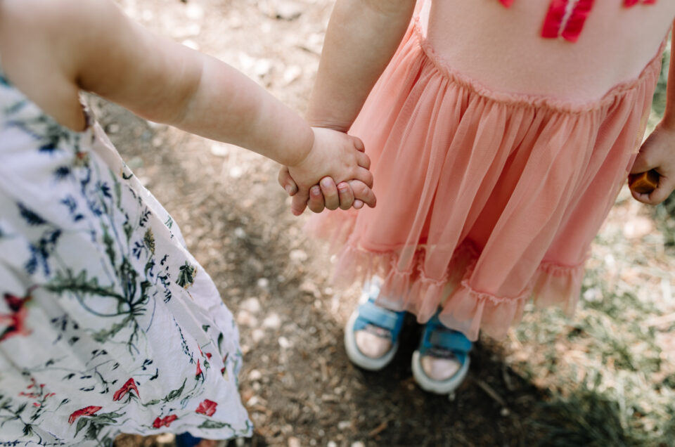 Two sisters are holding hands. One is wearing pink dress and blue trainers and another is wearing a white dress with colorful flowers. Family photographer in Basingstoke. Ewa Jones Photography