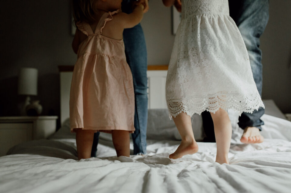 Family of four is jumping on a bed. One girl is wearing a white dress and another is wearing a pink dress. Family lifestyle photographer in Basingstoke. Ewa Jones Photography