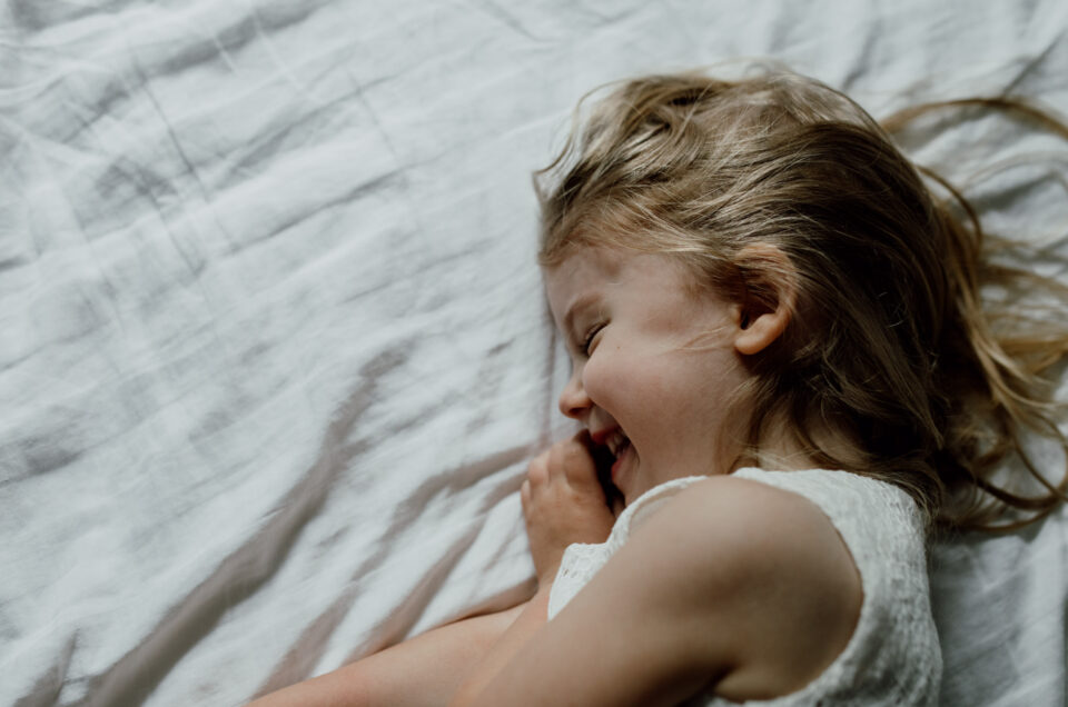 Girl is laying on bed and laughing. She is wearing a white dress. Candid moment. Child’s view on Covid-19. Family photographer in Basingstoke, Hampshire. Ewa Jones Photography