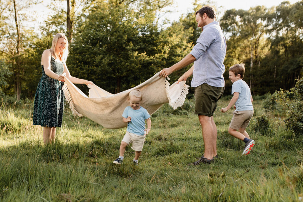 Mum and dad are holding blanket in the air and boys are running though the blanket. Family photo session during sunset. Lifestyle photography in Hampshire. Family photographer in Basingstoke. Ewa Jones Photography