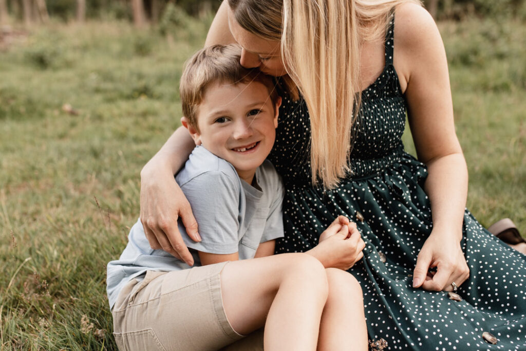 Mum and sun are sitting on the grass. Mum is kissing her boy on his head. Boy is wearing blue t-shirt and mum is wearing blue dress with white dots. Family photographer in Basingstoke. family photosession with Emma Reed. Ewa Jones Photography