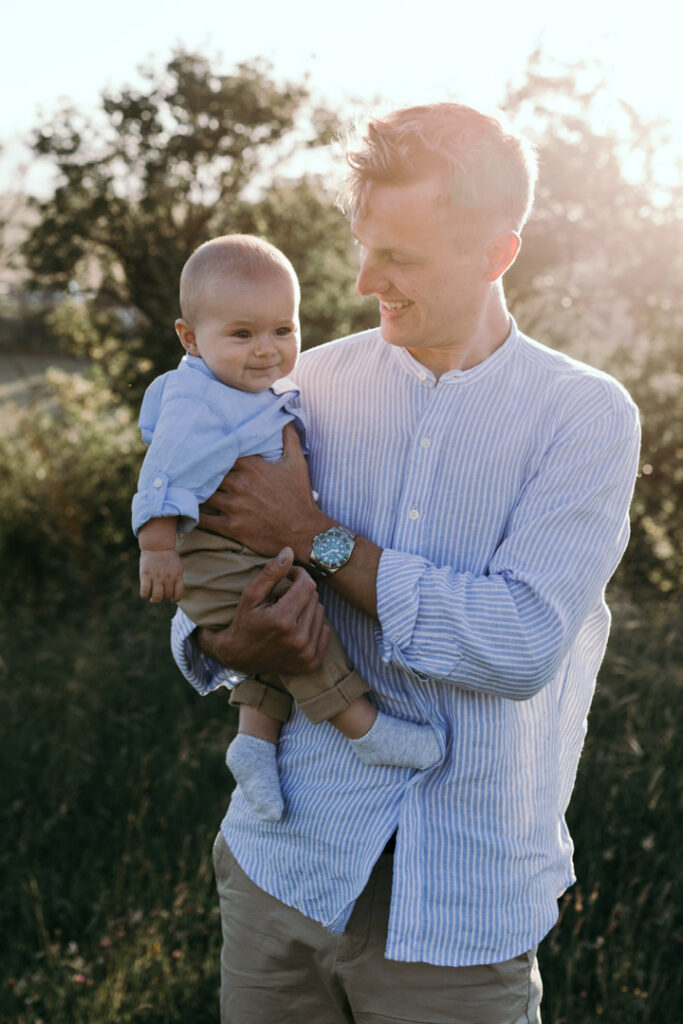 Dad is holding a little baby in his arms and looking at him. dad is wearing light blue shirt and boy is wearing blue shirt and light brown trousers. Dad is smiling to his baby. Candid photography. Lifestyle family session in Basingstoke. Ewa Jones Photography