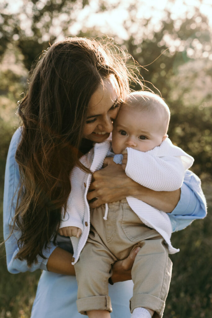 Mum is holding a baby and is smiling to her baby. Mum is standing with the sun shining on her back. Family photo session. Family photography in Basingstoke. Ewa Jones Photography