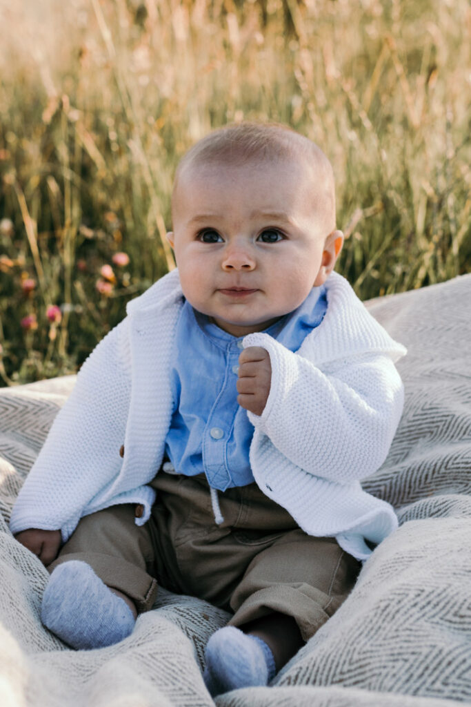 Boy is sitting on the blanket and looking at his mum. He is wearing a blue shirt, white cardigan and light brown trousers. Sun is shinning on his back. Candid photography. Family photographer in Hampshire. Ewa Jones Photography