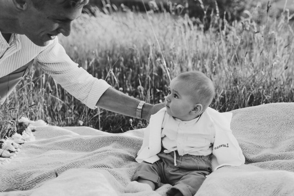 Little baby boy is sitting on the blanket and looking at his dad. His dad has a hand on his little boy shoulder and lovingly looking back at him. Black and white photograph.