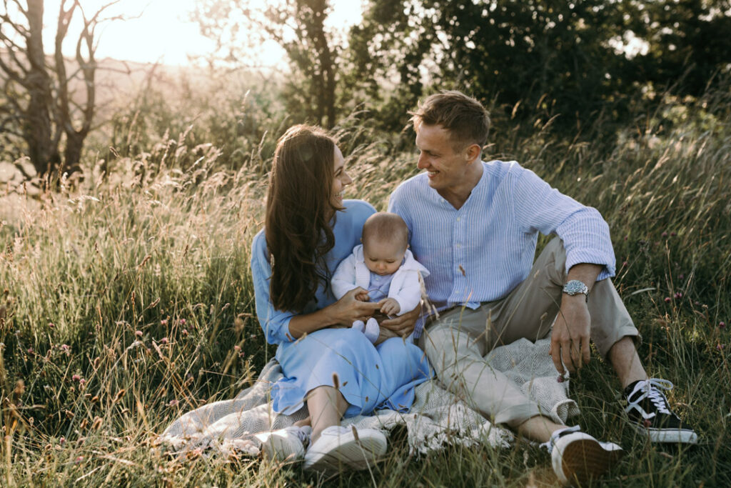 Mum and dad are sitting on the grass and looking at each other. Mum is holding a baby in her arms. family sunset photo session. Family photographer in Basingstoke. Ewa Jones Photography