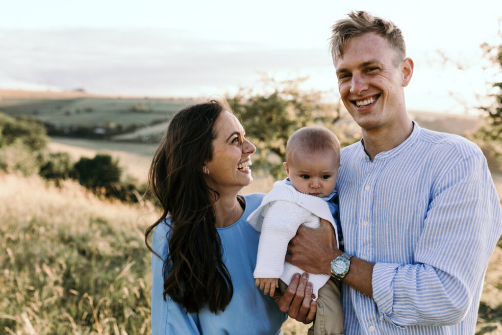 Mum and dad are standing and dad is holding a baby. Mum is looking at dad and smiling to him. Dad is also smiling. Mum is wearing blue dress and dad is wearing blue shirt. Happy family moment. Lifestyle family photo session. Family photography in Basingstoke. Ewa Jones Photography