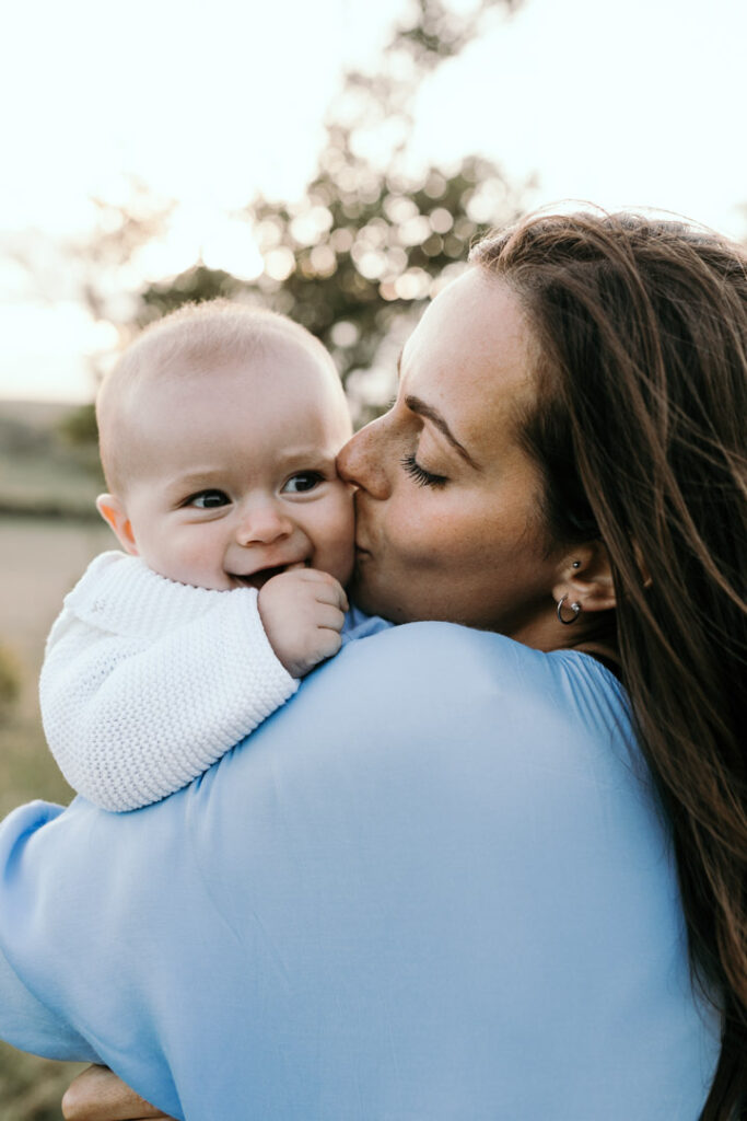 Sunset family photo session - Family photography - Ewa Jones Photography-36 Mum is holding and kissing a baby. Mum is wearing light blue dress and boy is wearing a white cardigan. Salisbury photo session in the hills.Lifestyle family session. Family photographer in Basingstoke. Ewa Jones Photography