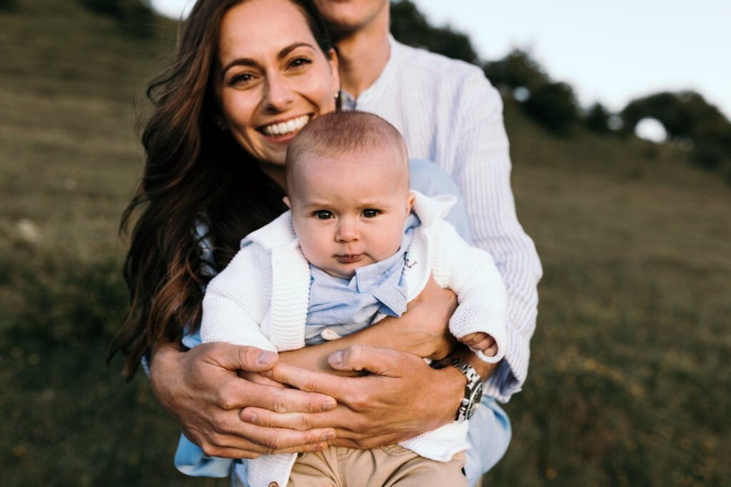 Mum and dad are holding a baby. Dad is standing behind mum wrapping his arms around mum and baby. Mum and baby are smiling. Lifestyle family session. Ewa Jones Photography