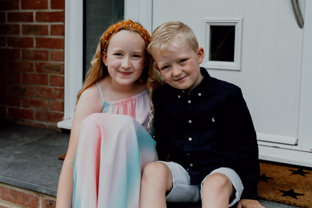 Brother and sister are sitting in front of their home and smiling. Family doorstep photo sessions in Hampshire. Ewa Jones Photography